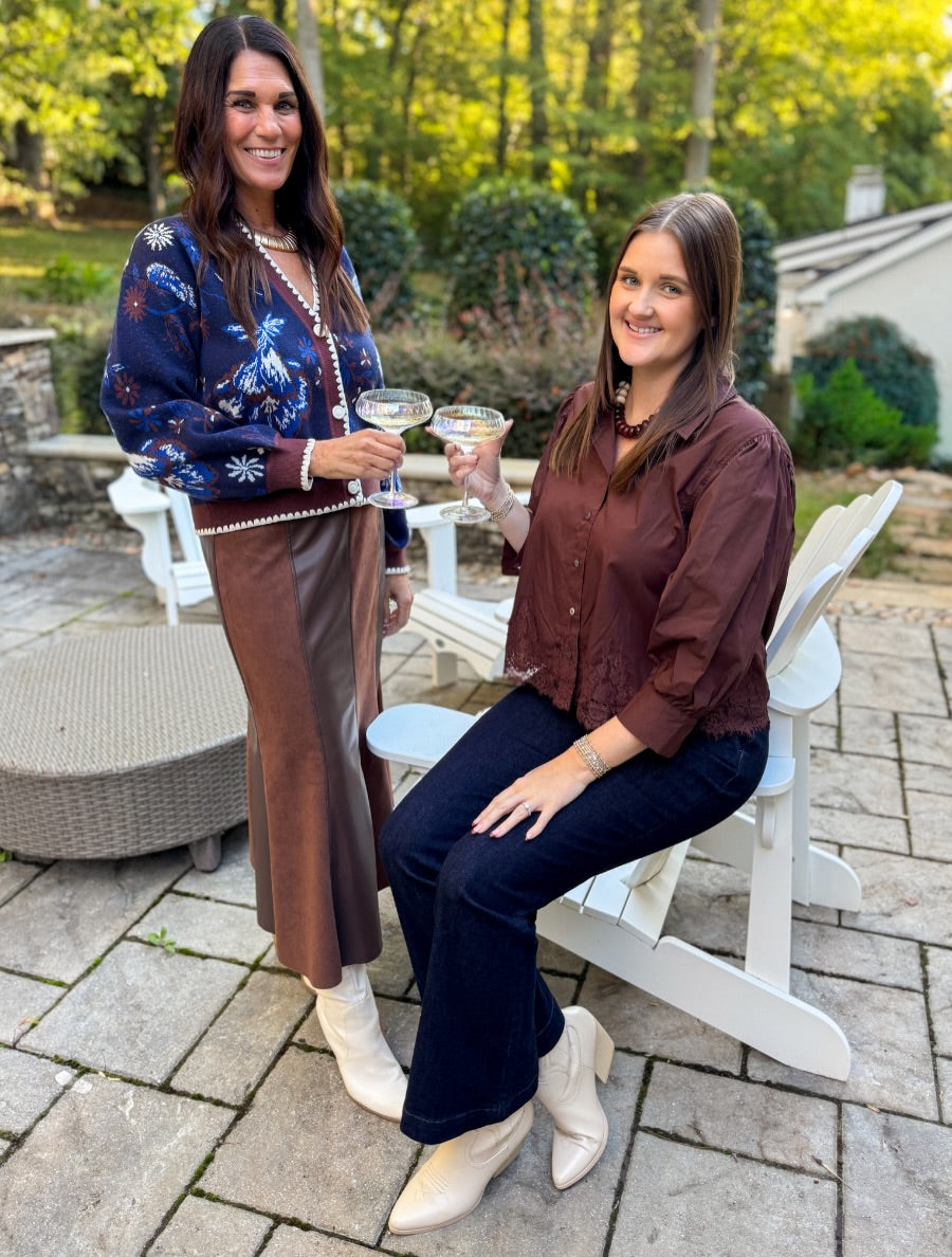 Two women posing on a stone patio outdoors. One stands wearing a navy blue floral cardigan, brown skirt, and white boots, holding a cocktail.