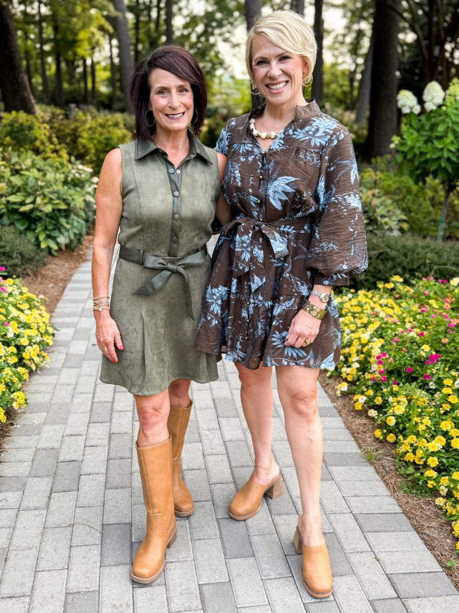 Two women standing on a paved garden path wearing short dresses and tan boots, with trees and flower beds in the background.