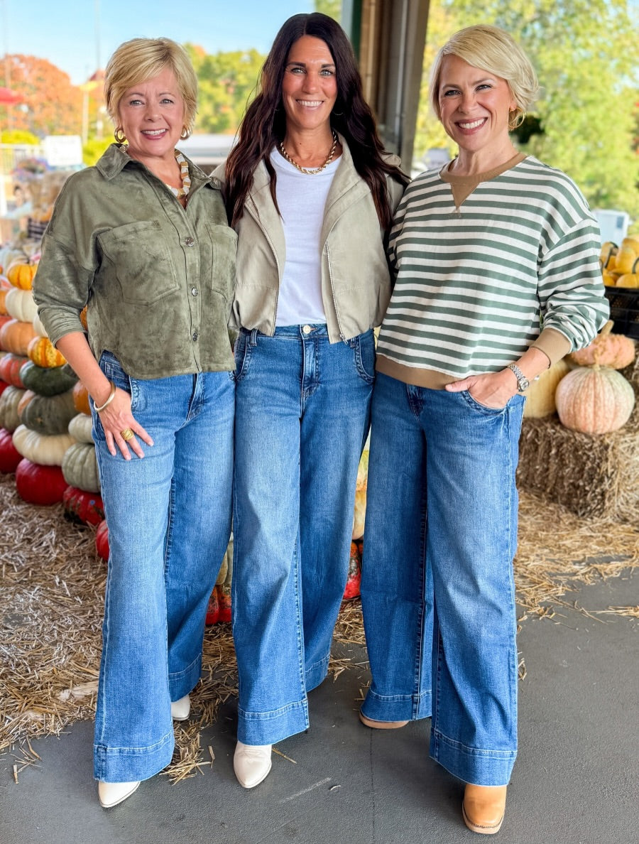 Three women standing together in front of a fall harvest display, wearing wide-leg blue jeans and different tops. The setting features hay bales and pumpkins.