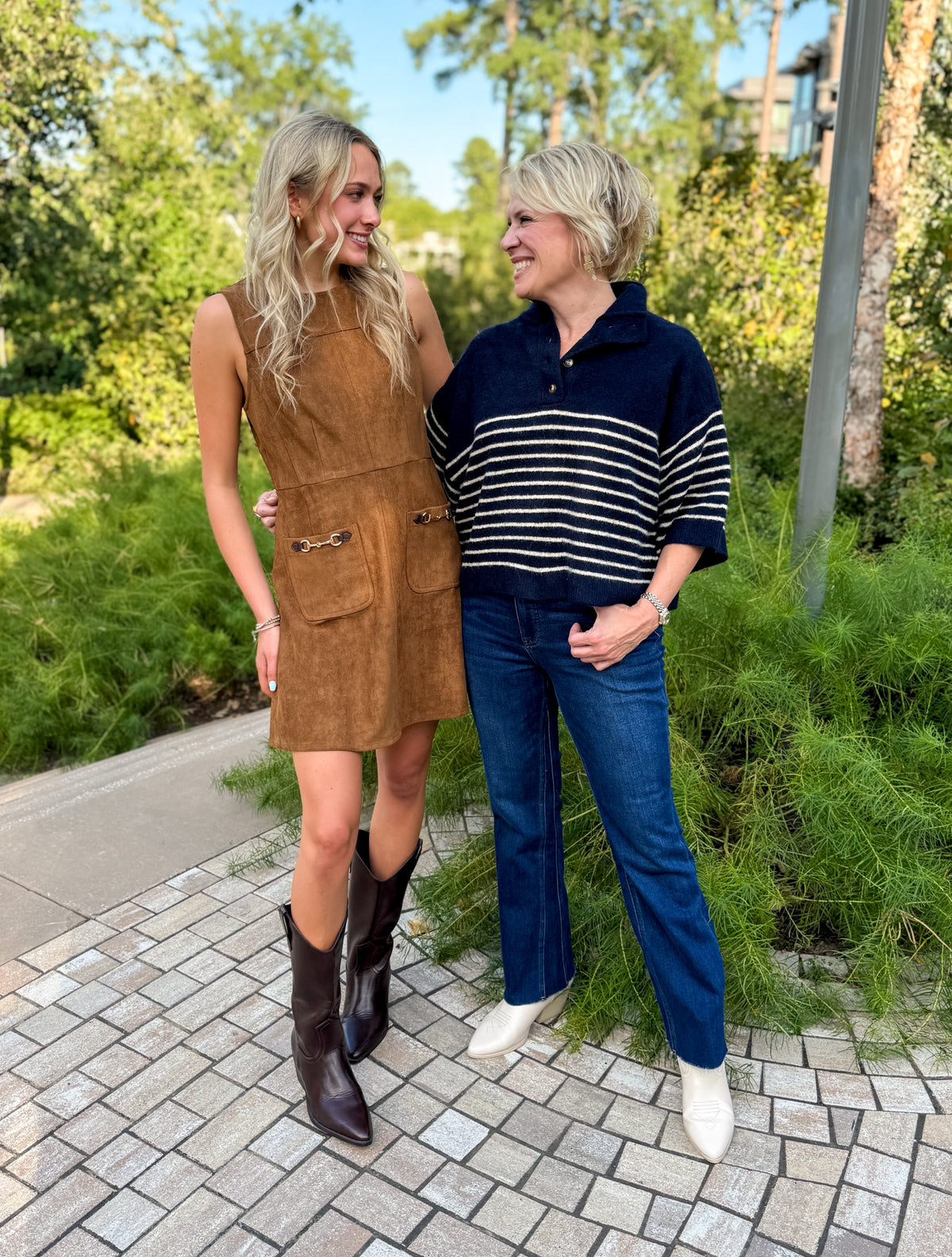 Two women standing outdoors on a paved walkway in a landscaped garden setting. The woman on the left wears a sleeveless brown suede-like mini dress with front patch pockets and dark brown pointed-toe boots.