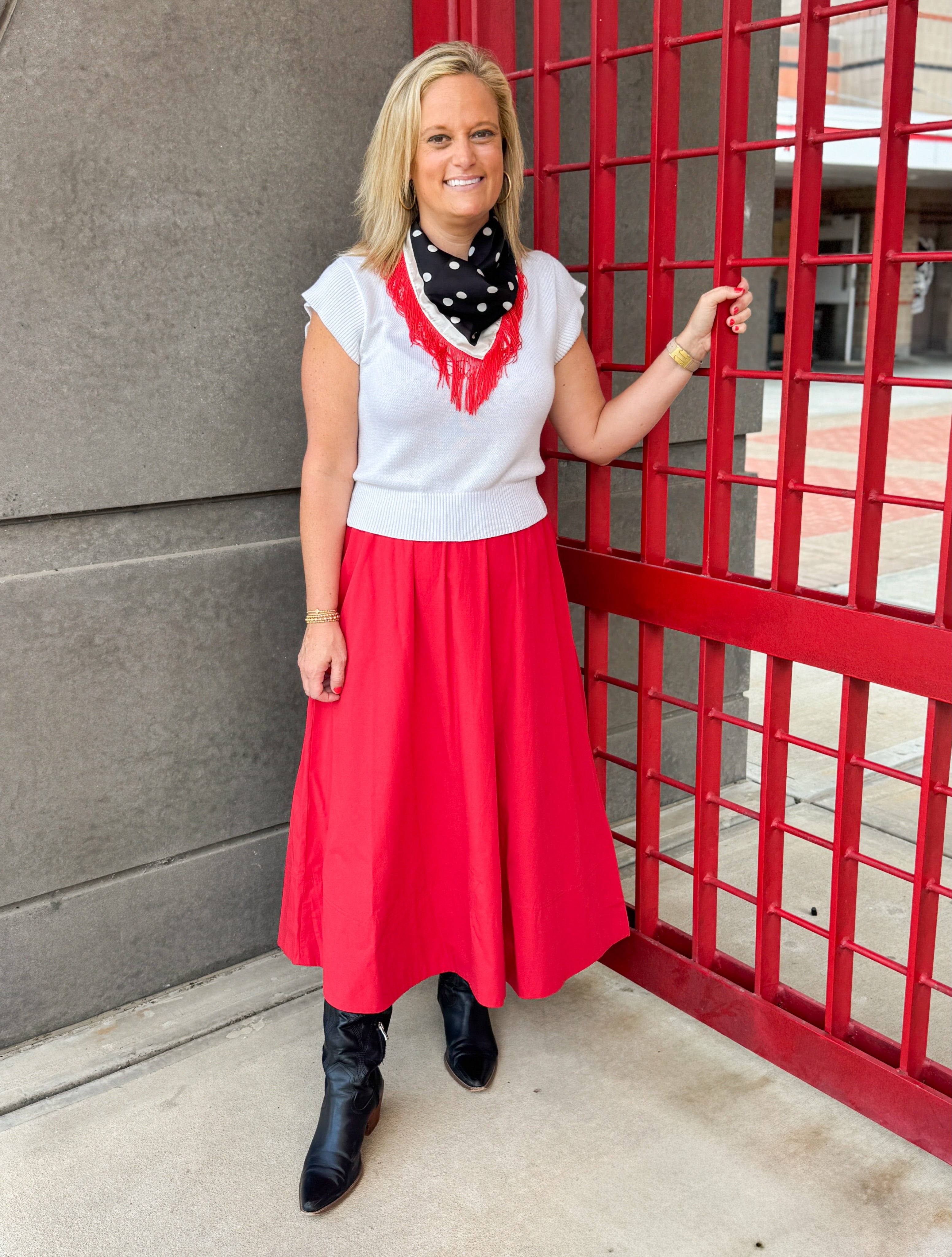 A woman stands outdoors next to a red metal gate, wearing a white sleeveless top with red fringe, a red midi skirt, a black and white polka dot bandana, and black ankle boots.