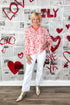 Woman in a pink blouse and white pants standing in front of a wall with red hearts and the word &