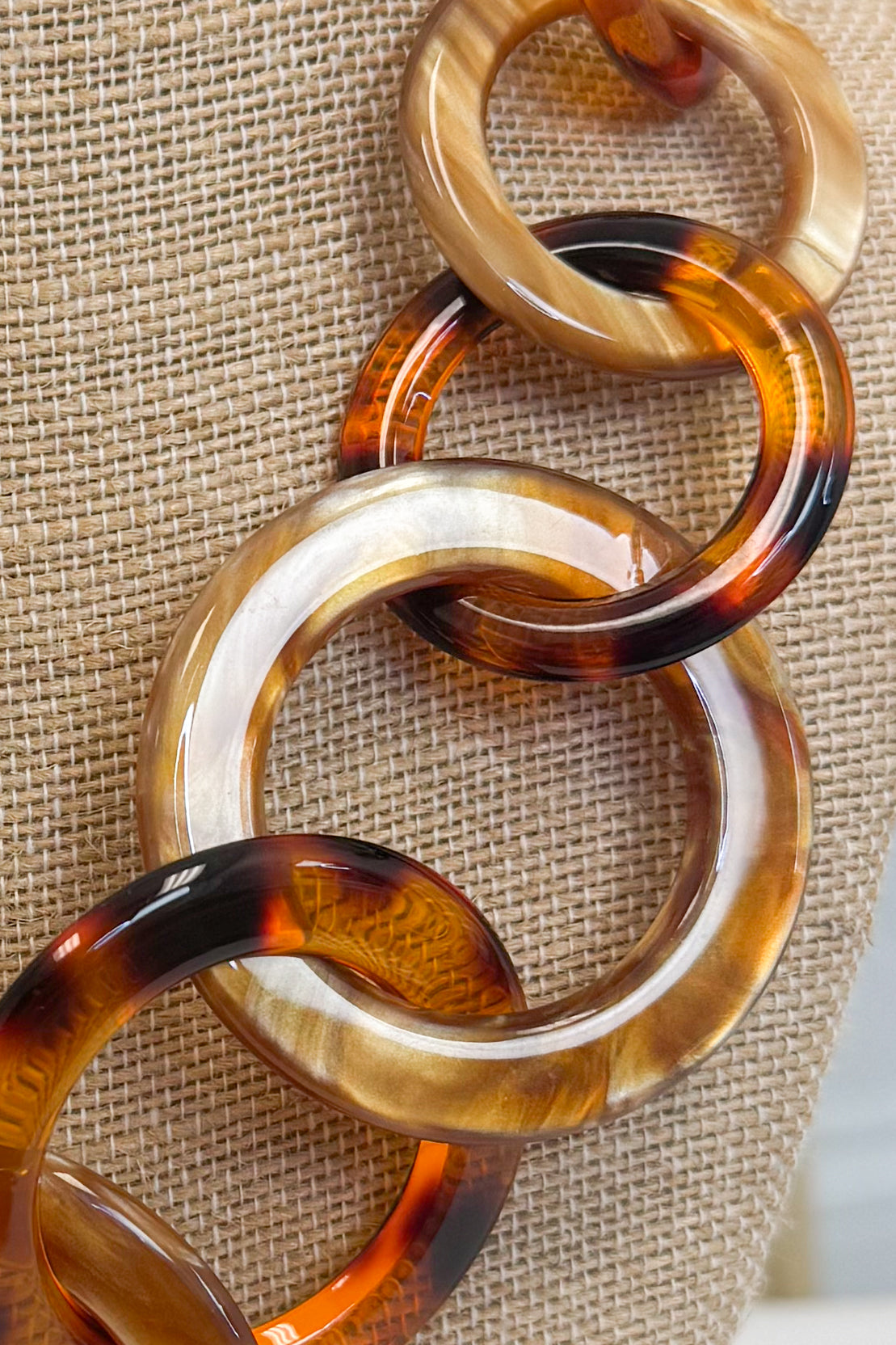 A close-up of a chunky chain-link necklace made of glossy tortoiseshell resin links in amber, brown, and cream tones, displayed on a textured beige fabric background.