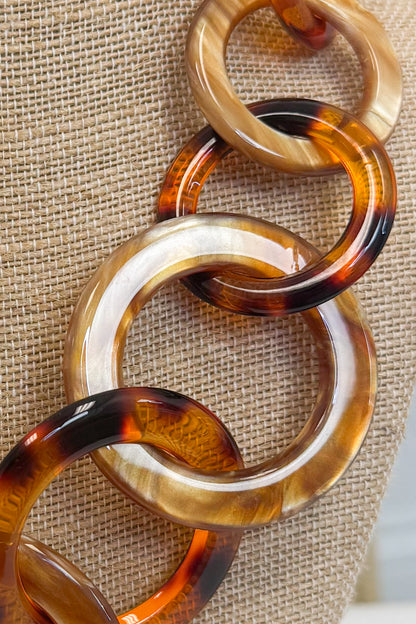 A close-up of a chunky chain-link necklace made of glossy tortoiseshell resin links in amber, brown, and cream tones, displayed on a textured beige fabric background.