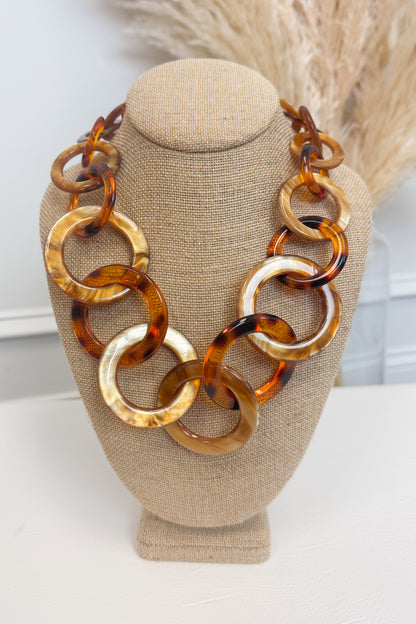 Chunky statement necklace made of large interlocking tortoiseshell and amber-colored links, displayed on a beige fabric jewelry bust against a light neutral background with dried grass decor.