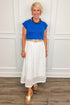 Woman wearing a blue top and white skirt with a wooden floor and white paneled wall background