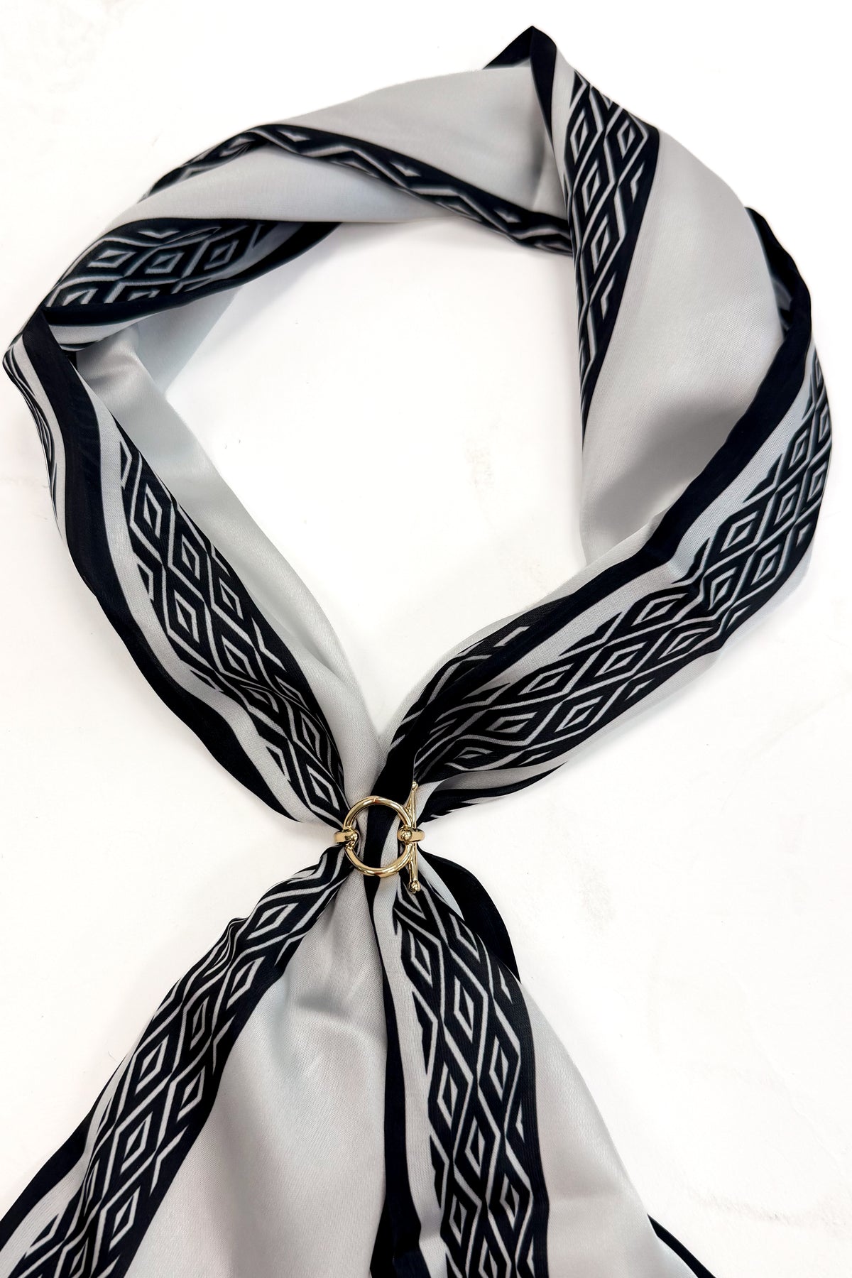 A lightweight scarf with a black and white geometric diamond pattern, tied in a knot with a small gold ring clasp, displayed on a clean white background.
