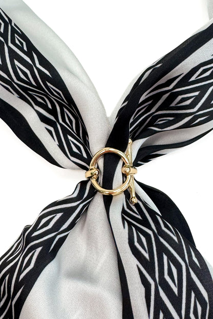 A close-up of a black and white patterned silk scarf tied in a knot with a gold ring clasp, set against a plain white background.