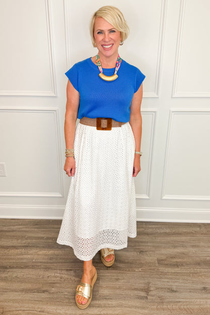Woman wearing a blue top and white skirt with a necklace, standing against a white paneled wall.