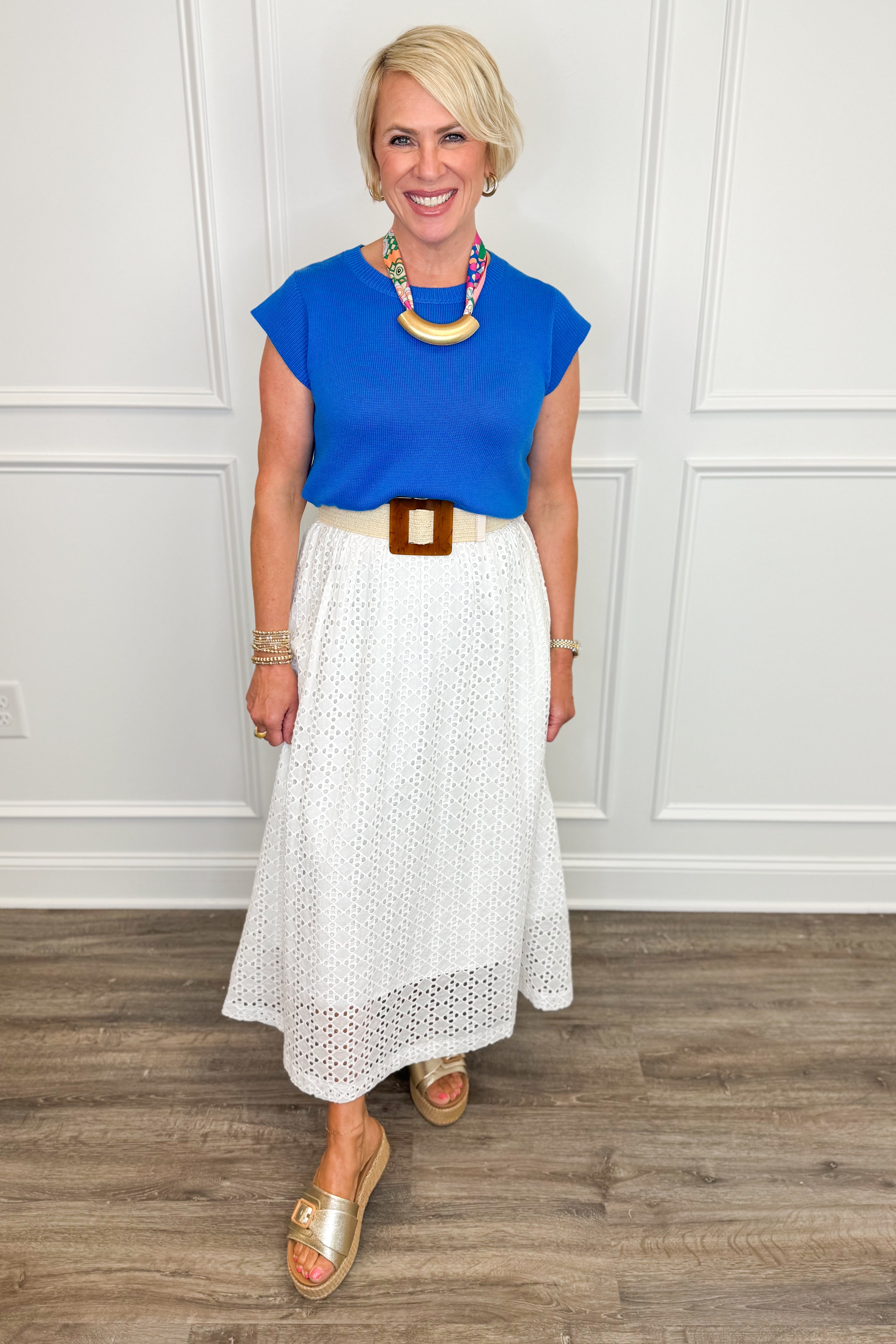 Woman wearing a blue top and white skirt with a wooden floor and white paneled wall background