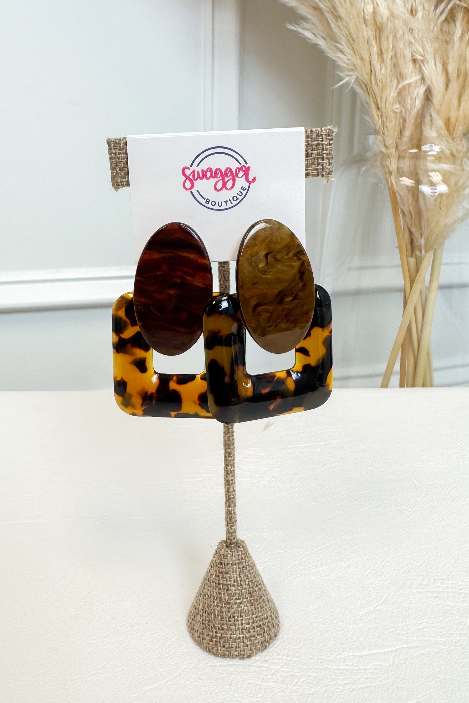 A pair of oval stud earrings with a tortoiseshell pattern, displayed on a stand with a card that reads &