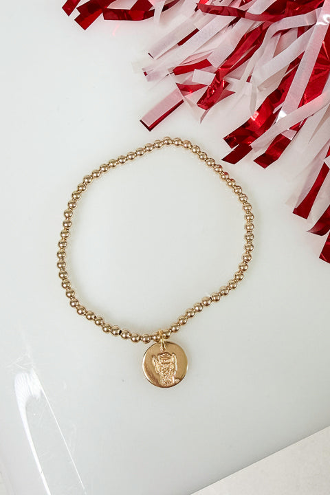 A gold beaded necklace with a circular pendant featuring a profile portrait, displayed on a white surface with decorative brown and white tinsel in the corner.
