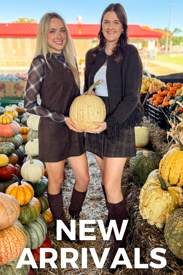 Two women standing in a pumpkin patch holding a large pale pumpkin, with a 'NEW ARRIVALS' text overlay at the bottom. The woman on the left wears a brown mini dress over a plaid long-sleeve top, and the woman on the right wears a dark brown fringed cardigan and matching plaid skirt.