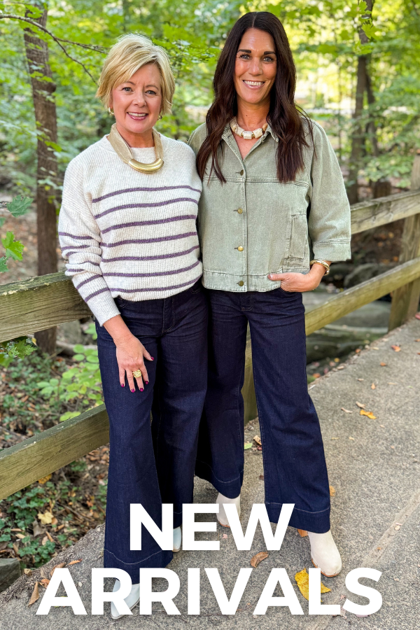 Two women smiling at the camera in a wooded outdoor setting, wearing casual fall outfits with sweaters, jackets, dark denim jeans, and neutral-colored shoes. White text reads “NEW ARRIVALS.”
