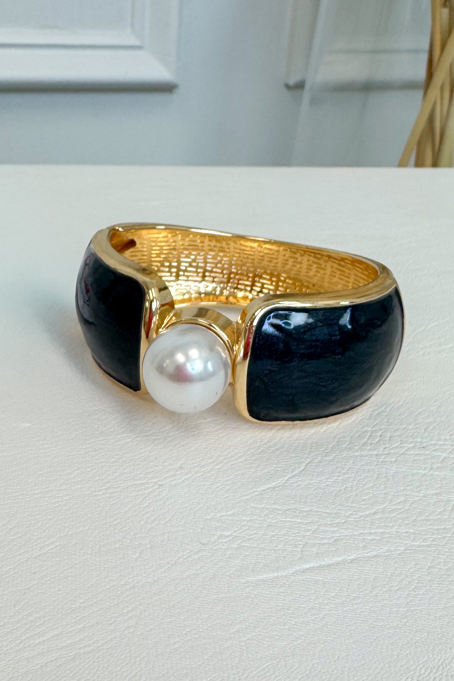 A gold-tone cuff bracelet featuring two glossy black enamel panels and a central white faux pearl, resting on a white textured surface.