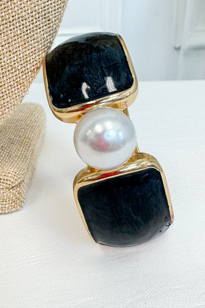 A gold-tone statement bracelet featuring two large square black stones and a central white pearl, displayed on a white surface next to a burlap jewelry stand.