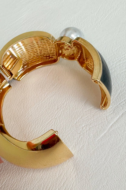 Gold-tone cuff bracelet with a textured interior, a black inlay, and a white pearl accent shown on a textured white background.