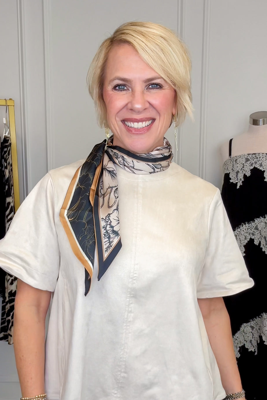 A smiling blonde woman wearing a cream-colored short-sleeve top and a patterned silk scarf with black, beige, and gold tones, standing in a boutique setting.