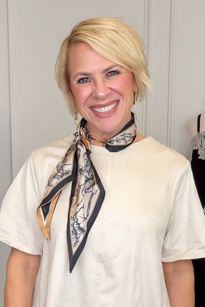 A smiling blonde woman wearing a cream short-sleeve top and a patterned silk neck scarf with black, beige, and gold details, posed against a light gray paneled wall.