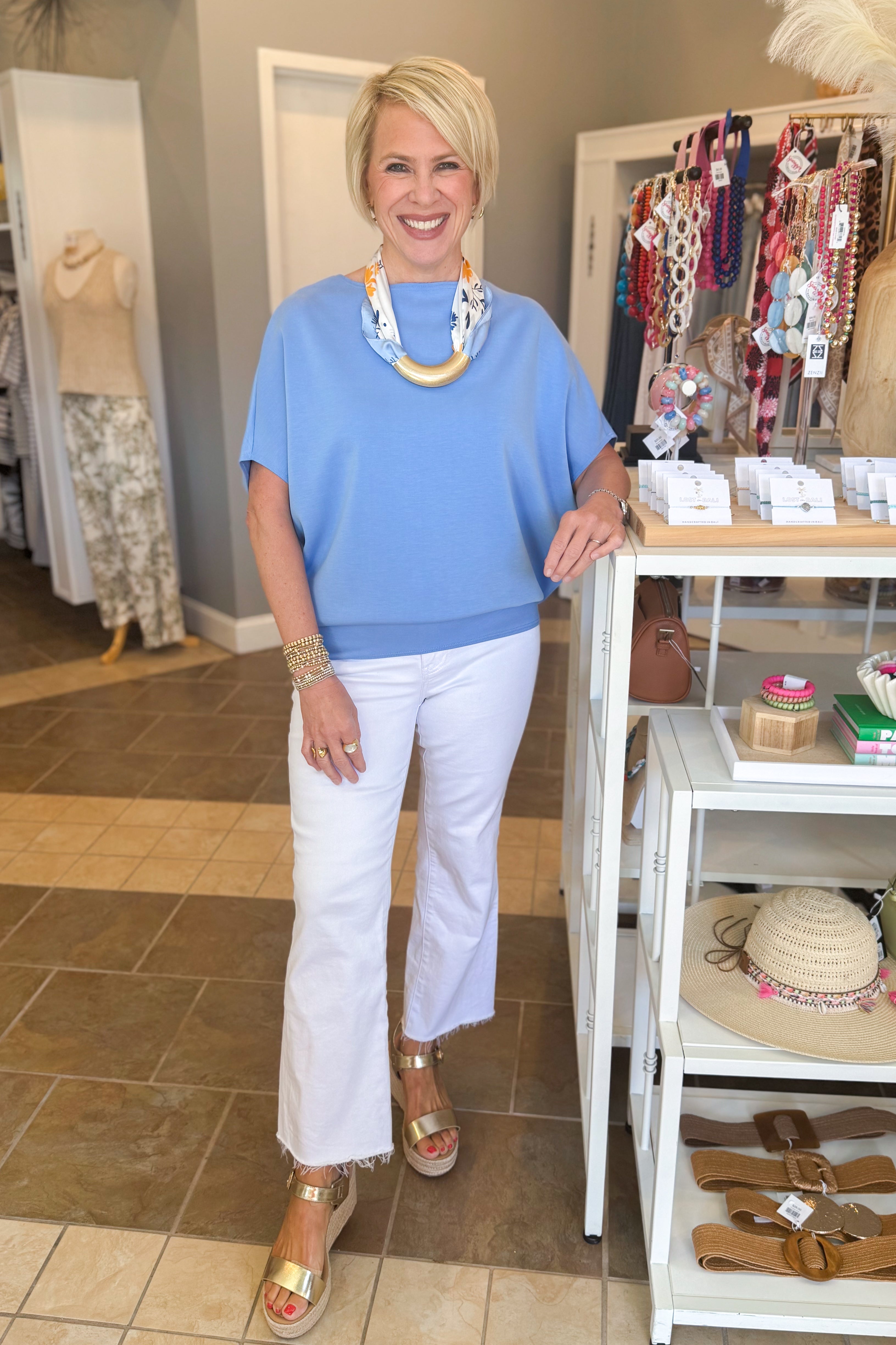 Woman in a blue top and white pants standing in a store with jewelry and clothing displayed.