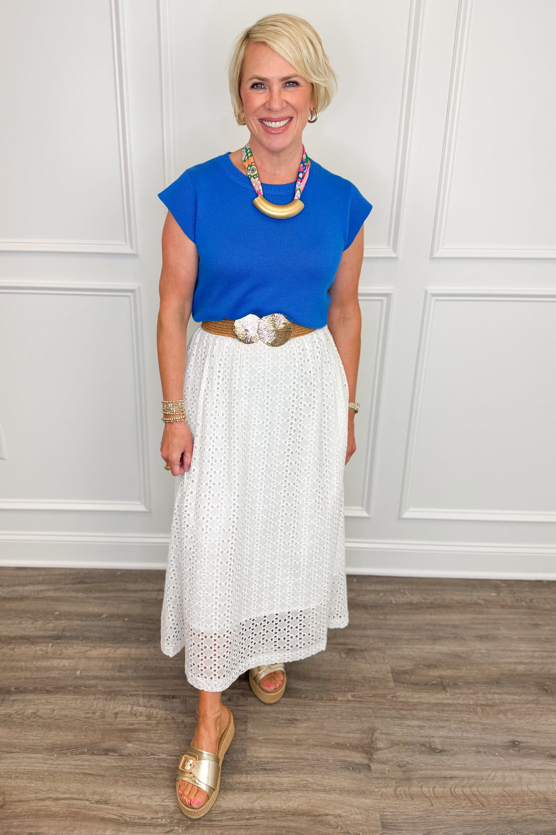 Woman wearing a blue top and white skirt with a necklace, standing against a white paneled wall.