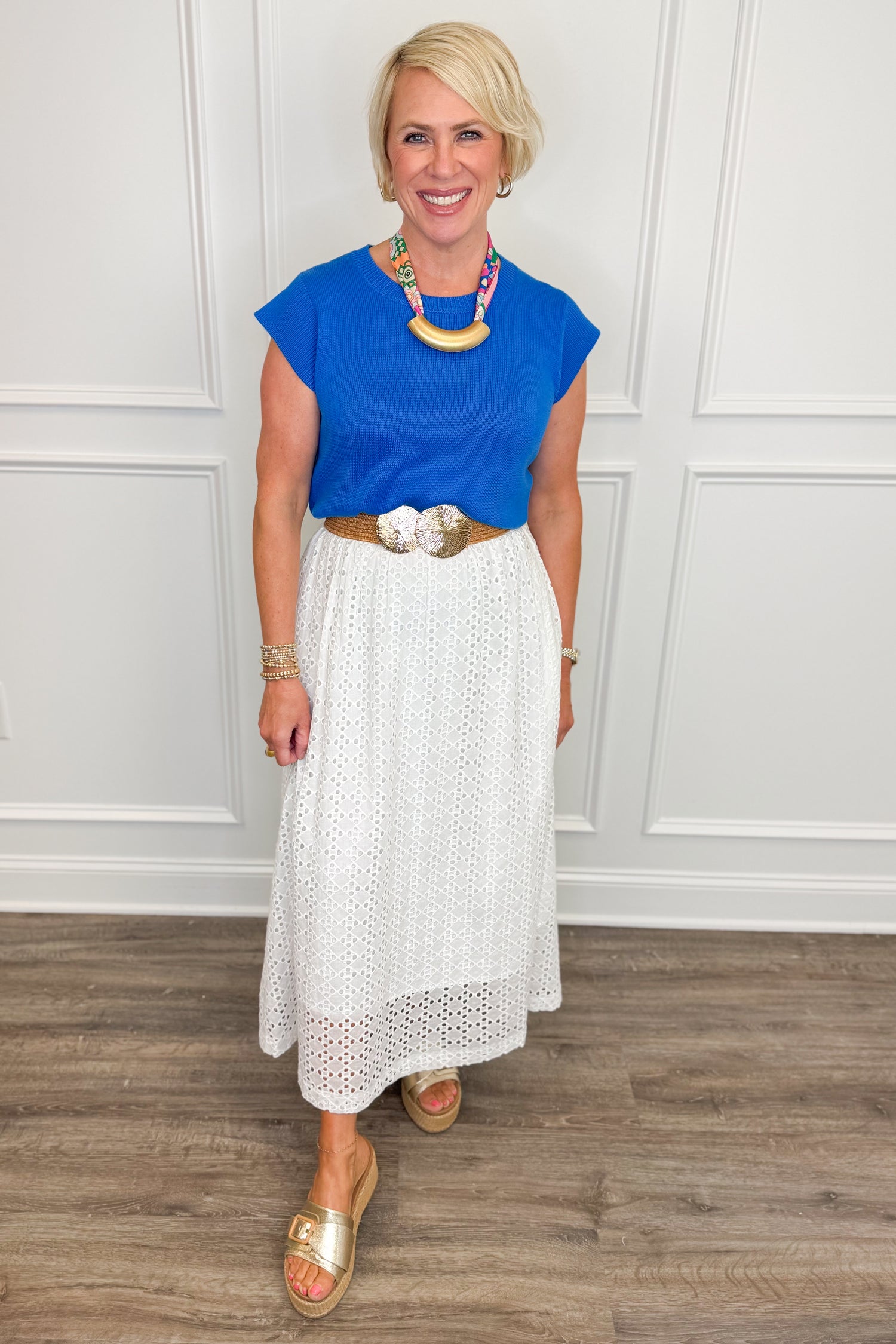 Woman wearing a blue top and white skirt with a necklace, standing against a white paneled wall.