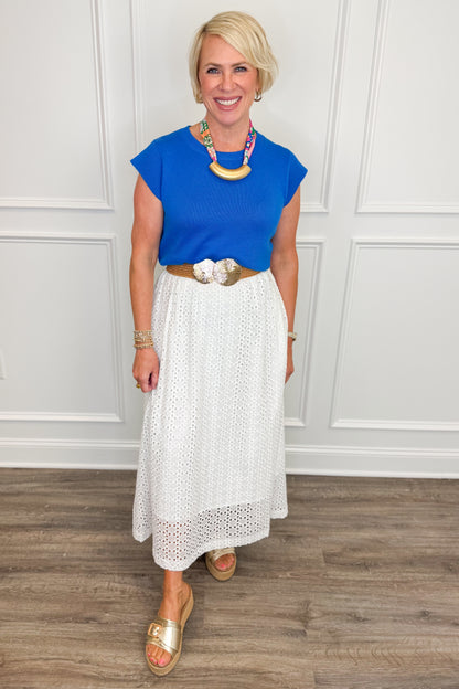 Woman wearing a blue top and white skirt with a necklace, standing against a white paneled wall.