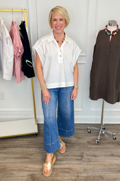 Woman in a white blouse and blue jeans standing in a clothing store with racks and mannequins in the background.