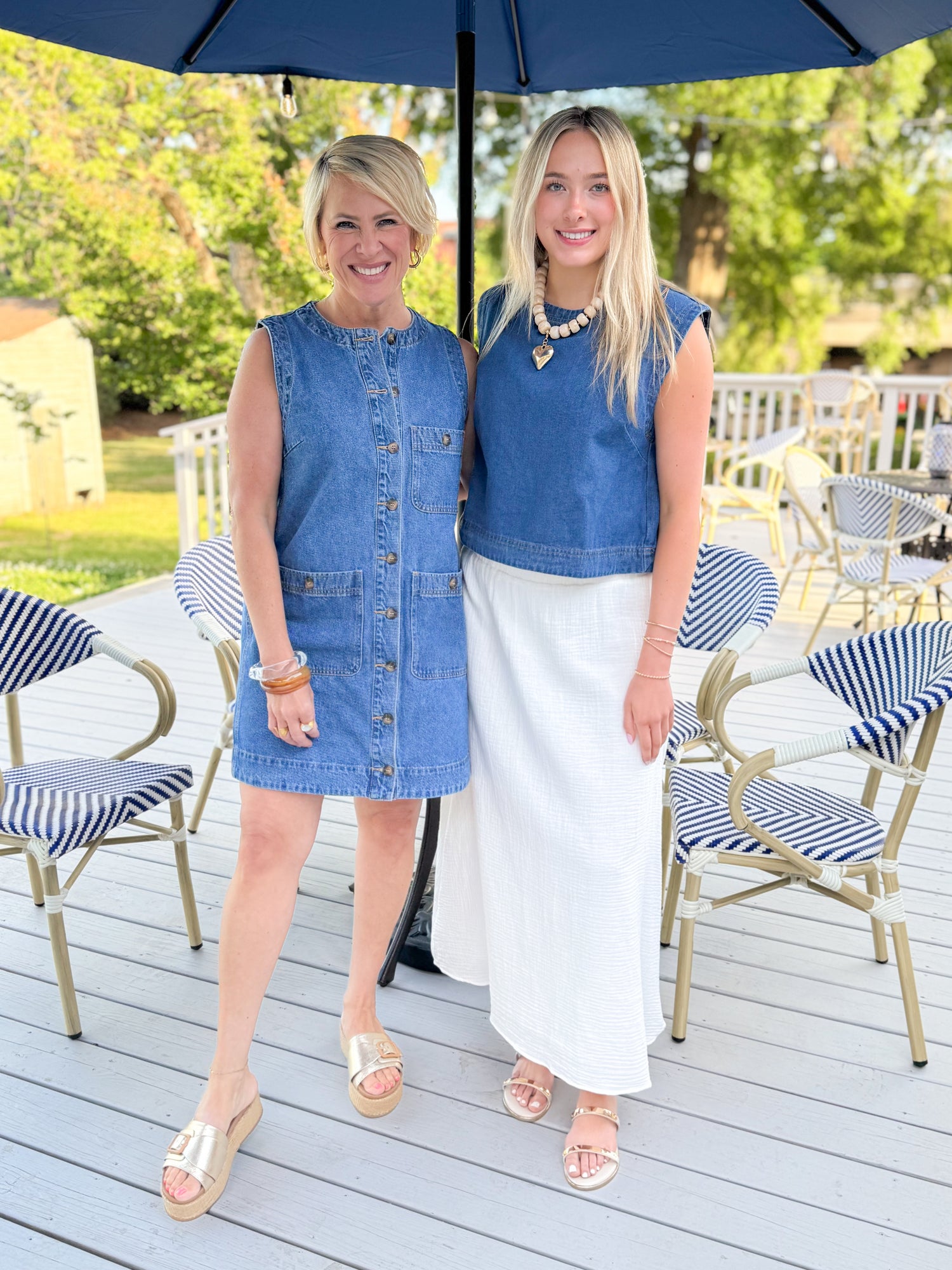 Two women in matching blue sleeveless dresses standing under an umbrella on a patio.