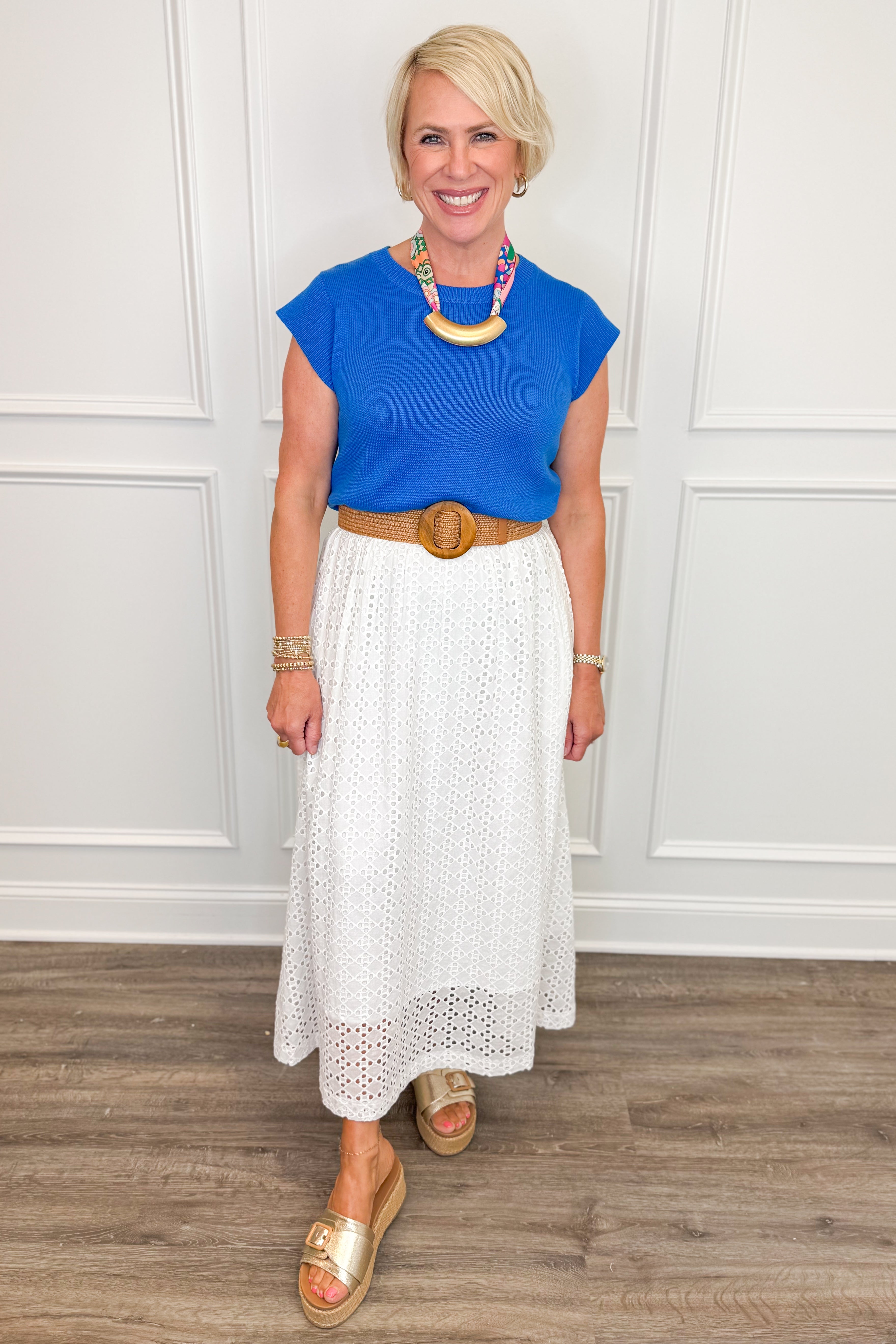 Woman wearing a blue top and white skirt with a wooden circle belt, standing against a white paneled wall.