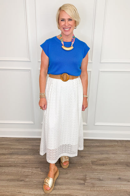 Woman wearing a blue top and white skirt with a wooden circle belt, standing against a white paneled wall.